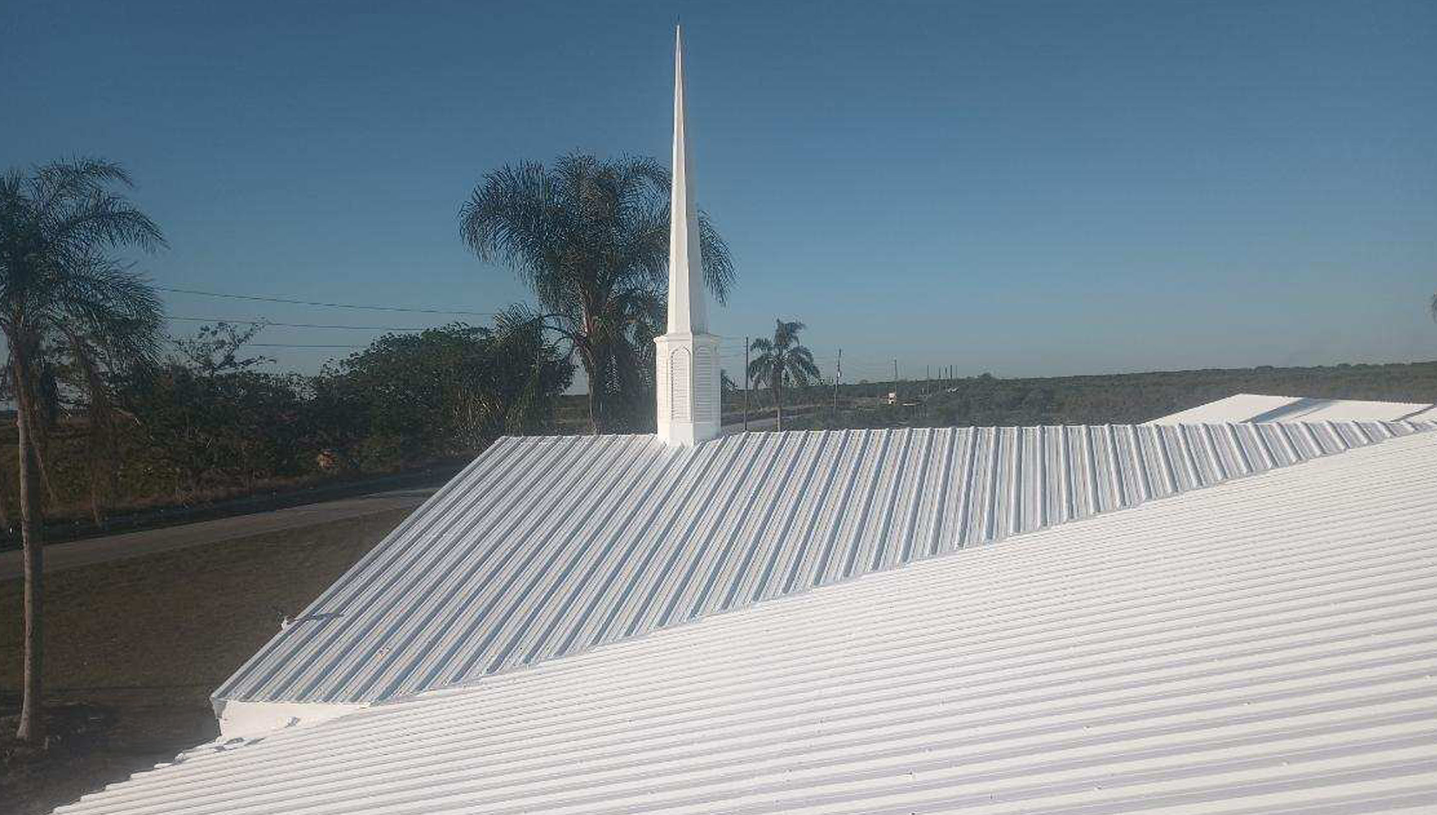 A white metal church roof with a tall white spire against a clear blue sky.