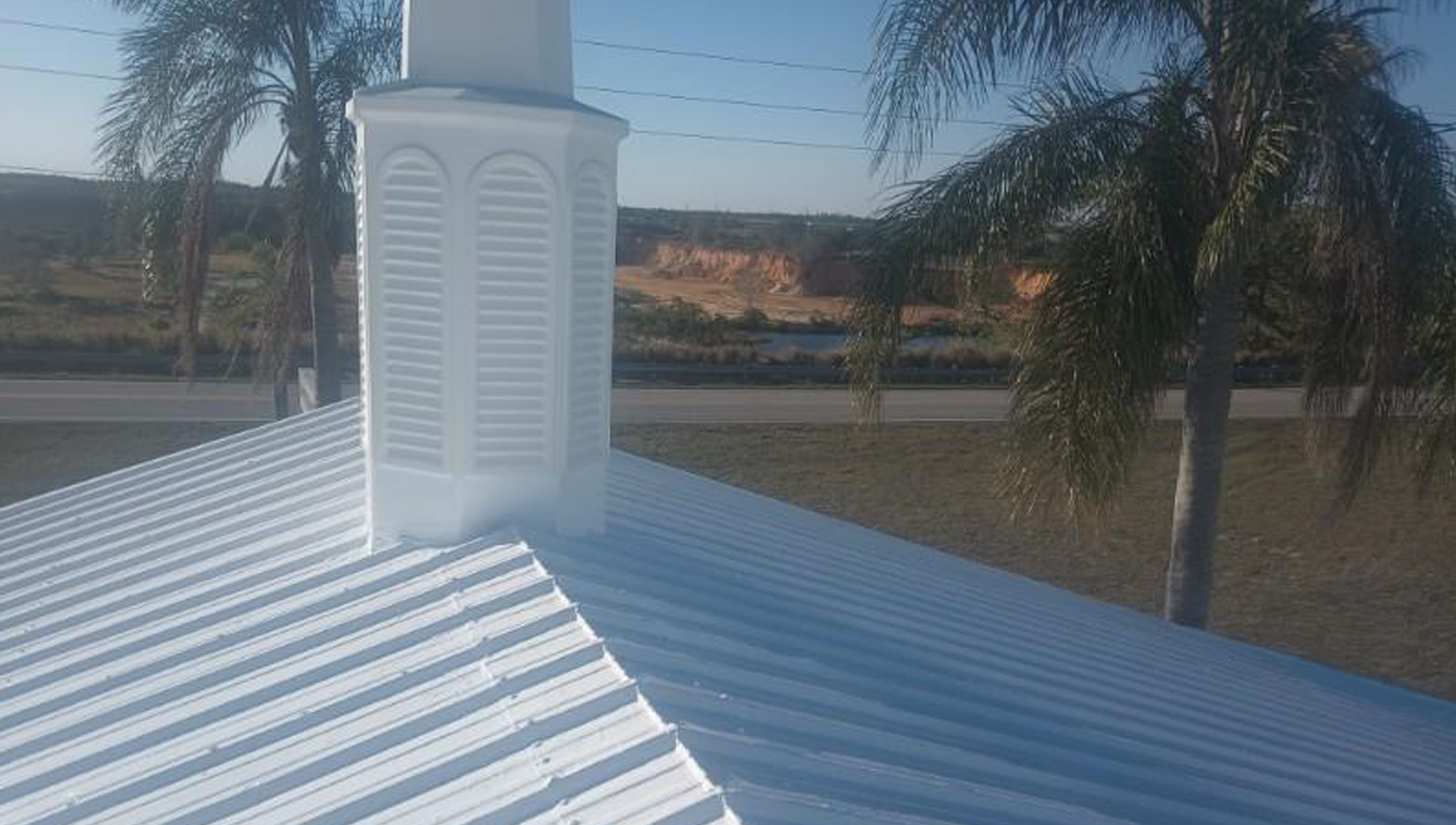 A church's white metal roof with a louvered cupola and palm trees in the background.